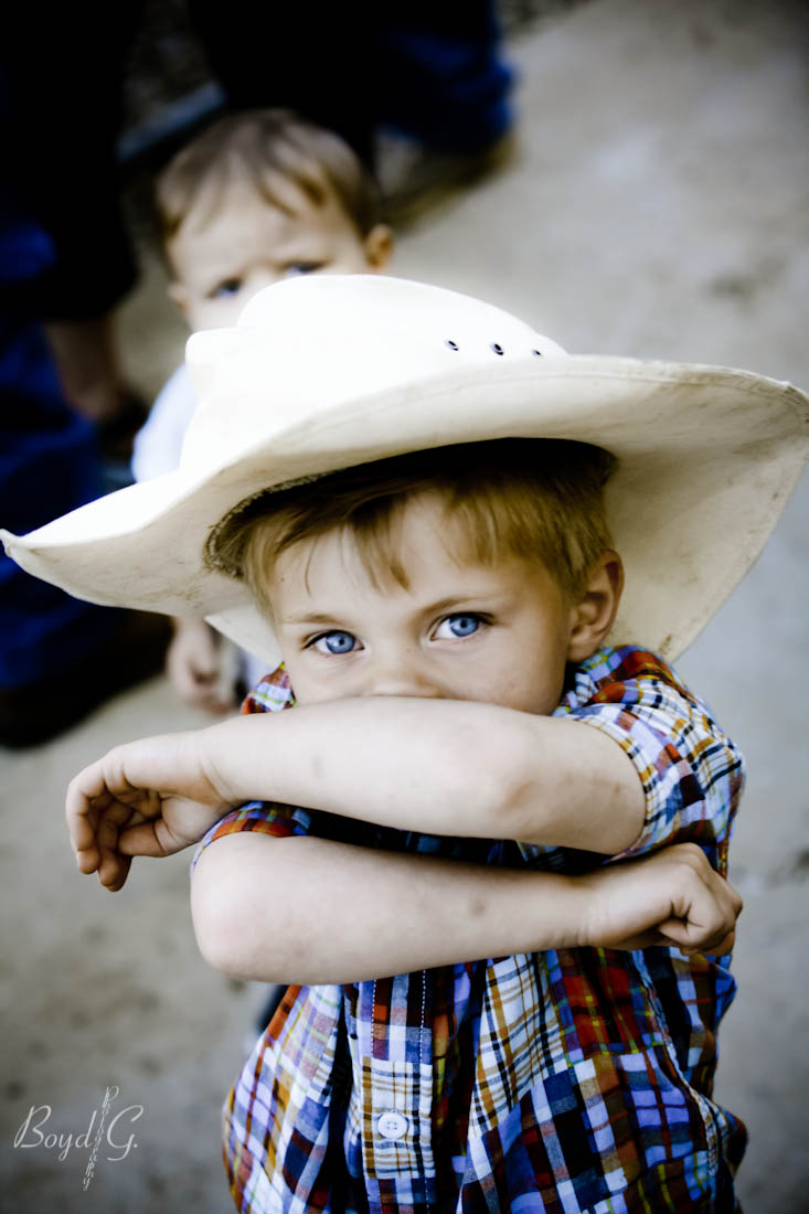 Little boy with in a cowboy hat covers his face and looks up inquisitively at the camera at a rural wedding.