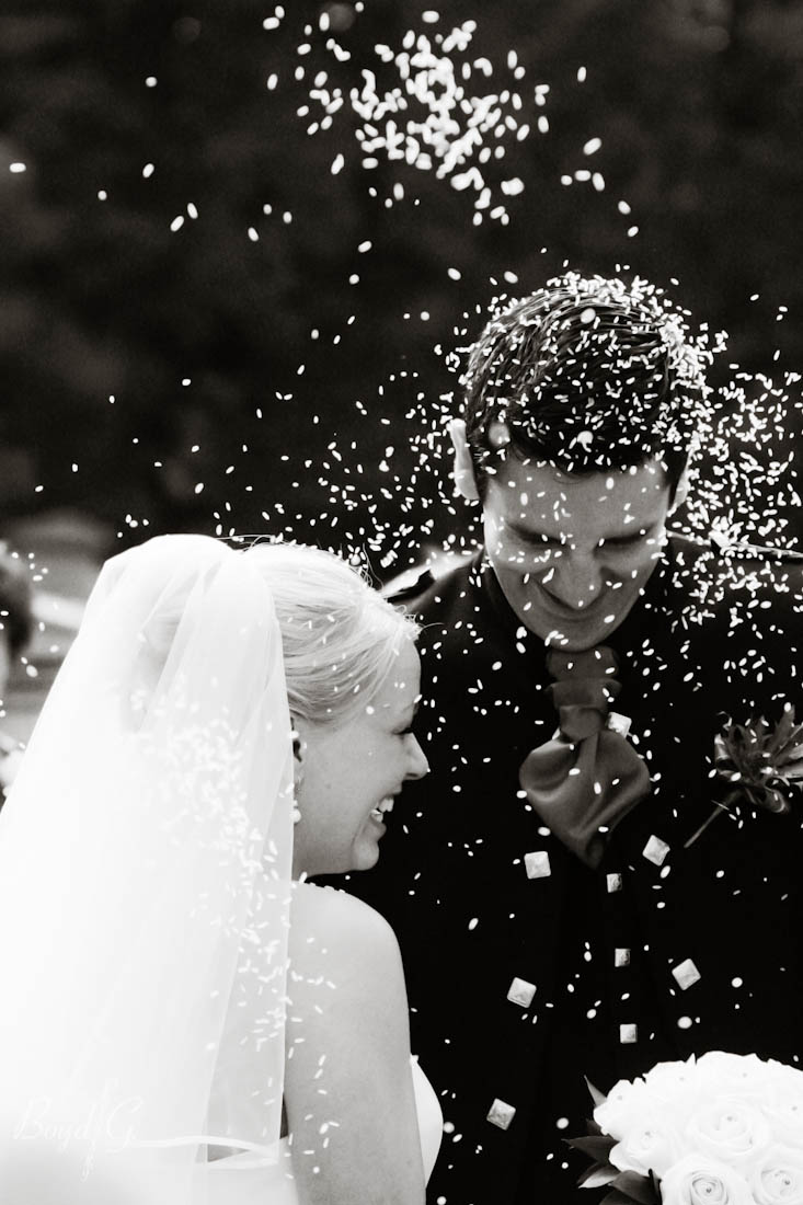 Bride and groom smile emphatically and cringe and rice is being thrown at them by their guests.